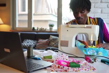 While learning to stitch on a sewing machine, a man is watching a video on his laptop for a training course, on a table covered with pretty materials and cotton spools